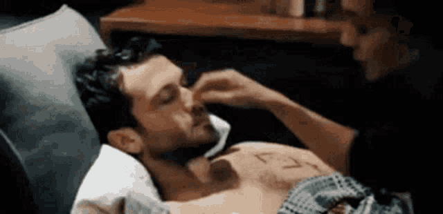 a man without a shirt is laying in a bed with a woman touching his face .