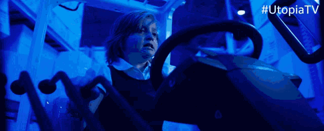 a woman is driving a car in a dark room with # utopiatv on the bottom right