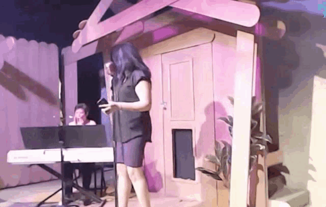 a woman singing into a microphone while standing in front of a cardboard house