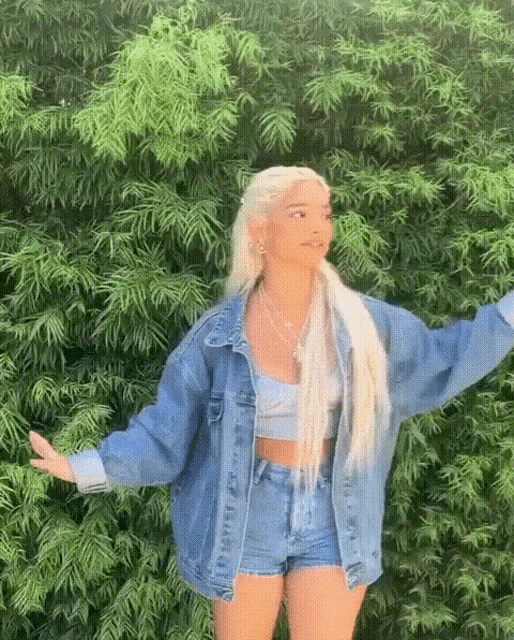 a blonde woman wearing a denim jacket and shorts is standing in front of a green bush .