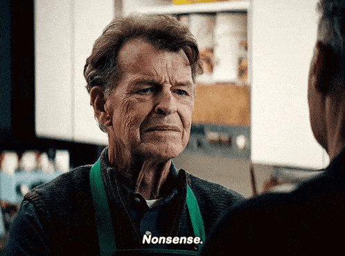 an older man wearing a green apron says nonsense to another man