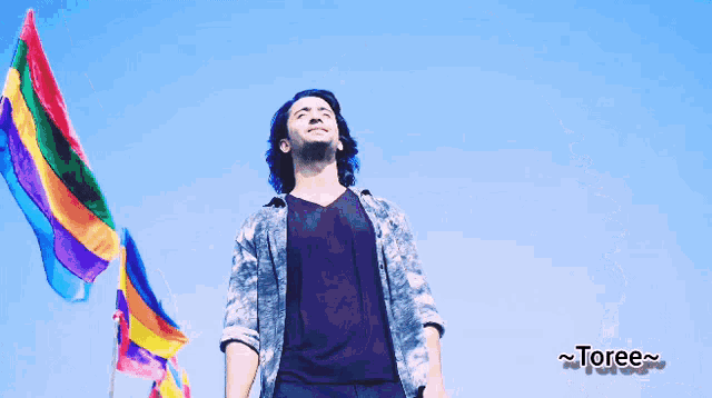 a man stands in front of a blue sky with rainbow flags behind him and the word toree on the bottom