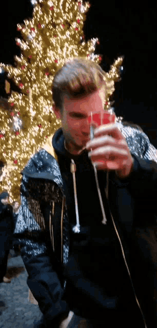 a man in a sequined jacket holds a glass in front of a christmas tree
