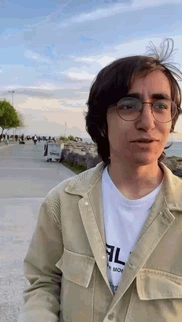 a young man wearing glasses and a tan jacket is standing in front of a body of water .