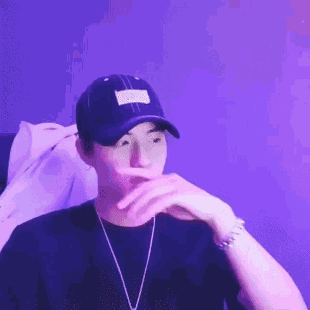 a young man wearing a hat and a necklace is covering his mouth with his hand .