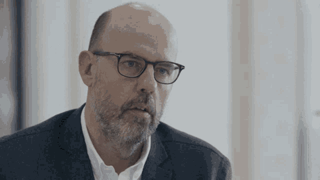 a bald man with glasses and a beard wears a suit