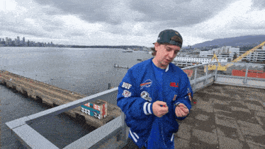 a man wearing a blue buffalo bills jacket stands on a balcony overlooking a body of water