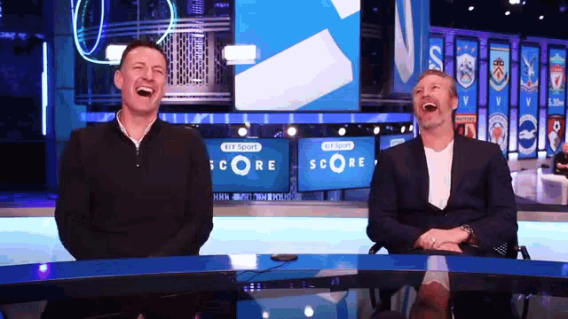 two men are laughing in front of a score television screen