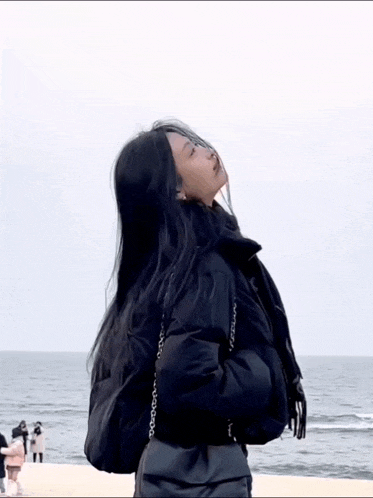 a woman in a black jacket is standing on a beach looking up at the sky .