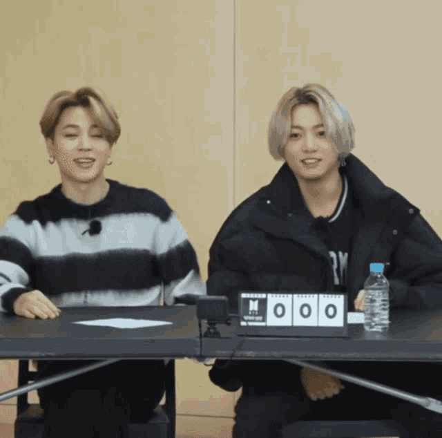 two young men sit at a table with a bottle of water and a sign that says ' bts ' on it