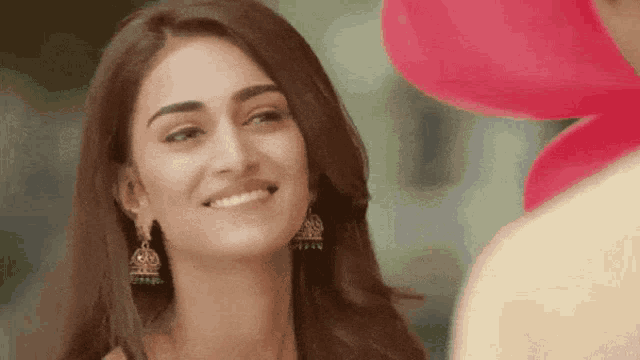 a woman wearing earrings is smiling in front of a pink flower