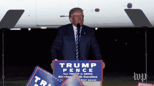 donald trump stands at a podium holding a sign that says trump pence