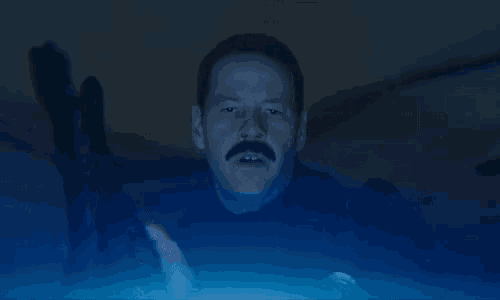a man with a mustache is sitting in a dark room with a blue light shining on him .