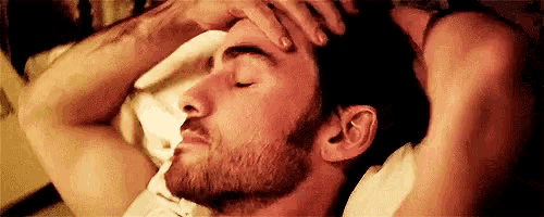 a man with a beard is laying in bed with his eyes closed and holding his head .
