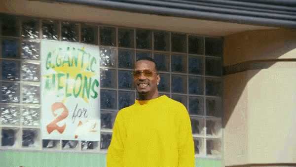 a man in a yellow sweater stands in front of a sign that says elegant 2 melons