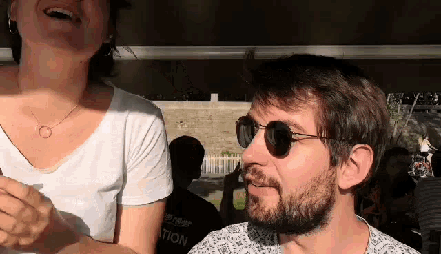 a man with a beard and sunglasses is laughing next to a woman in a white shirt .