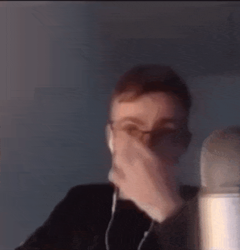 a man wearing glasses and ear buds is covering his mouth with his hand .