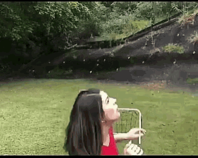 a woman in a red shirt is blowing soap bubbles in a park while looking up at the sky .