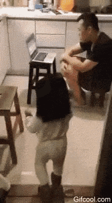 a little girl is standing in front of a laptop computer while a man sits on a stool in a kitchen .