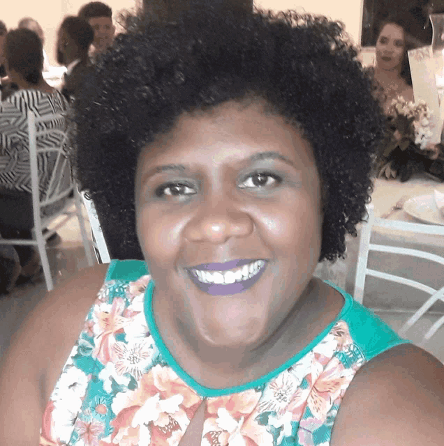 a woman wearing purple lipstick and a floral shirt smiles for the camera