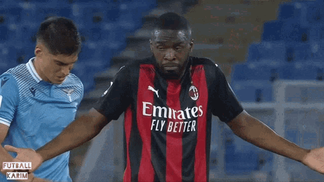 a soccer player wearing an emirates fly better jersey stands next to another player