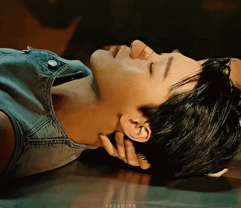 a woman in a denim vest is laying on the floor with her eyes closed and her hand on her neck .