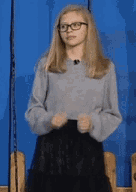 a young girl wearing glasses and a gray sweater is standing in front of a blue curtain .
