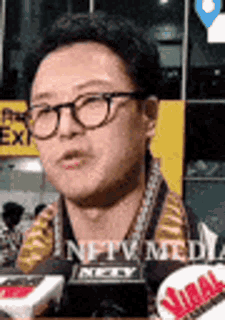 a man wearing glasses is talking into a microphone in front of a yellow sign .