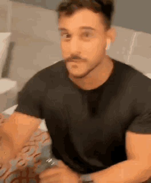 a man with a mustache is sitting on a bed wearing a black shirt .