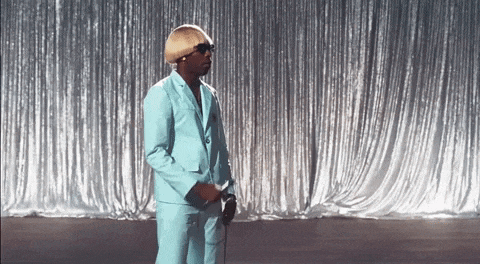 a man in a light blue suit and sunglasses is standing in front of a silver curtain .