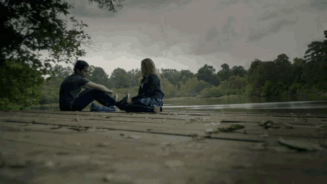 a man and a woman sit on a wooden dock overlooking a lake