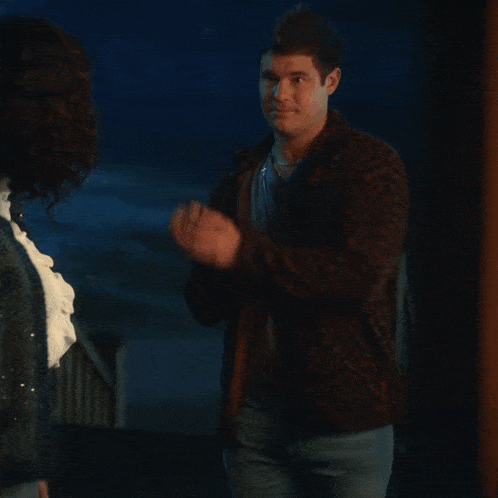 a woman in a blue cardigan stands in front of a man in a brown jacket