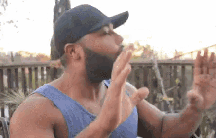 a man with a beard wearing a baseball cap and a blue tank top is eating a hot dog .