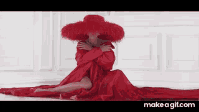 a woman in a long red dress and red hat is sitting in front of a white wall .