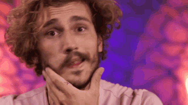 a man with curly hair and a beard is making a funny face while holding his hand to his chin .