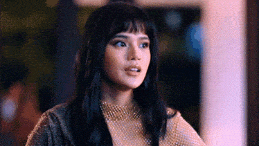 a woman with long black hair and bangs is wearing a gold top and necklace