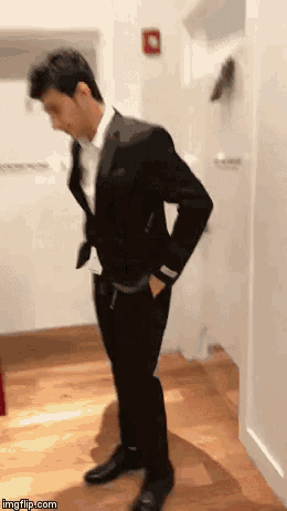a man in a black suit is standing on a wooden floor with his hands in his pockets .