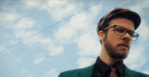 a man wearing glasses and a hat is standing in front of a cloudy sky
