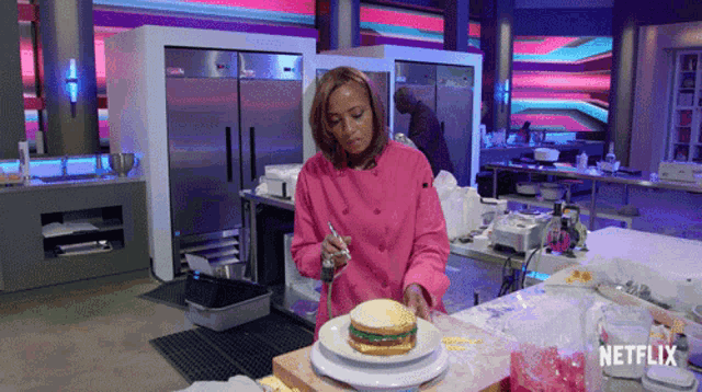 a woman in a pink chef 's jacket is decorating a sandwich on a plate in a kitchen with a netflix logo