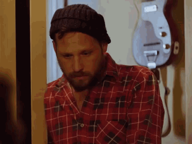 a man wearing a plaid shirt and a beanie stands in front of a guitar hanging on the wall