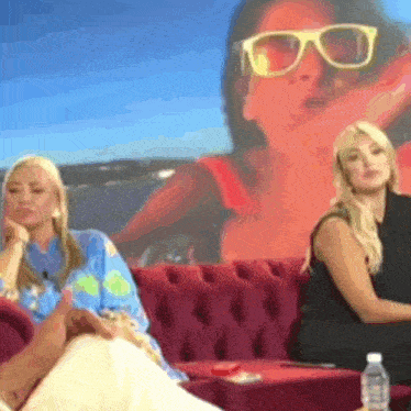 two women are sitting on a red couch in front of a wall with a picture of a woman wearing sunglasses
