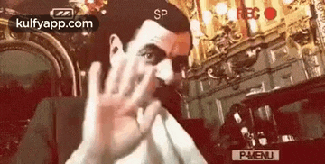 mr bean is making a funny face while sitting in a chair and covering his face with his hand .