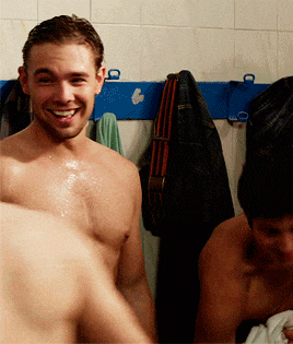 a shirtless man smiles in a locker room with other men