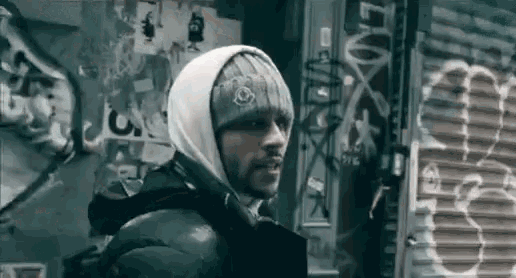 a man wearing a hooded jacket and a beanie is standing in front of a wall with graffiti on it .