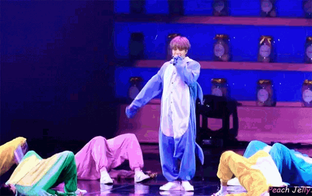 a man in a blue shark costume is singing into a microphone while a group of dancers perform on stage .