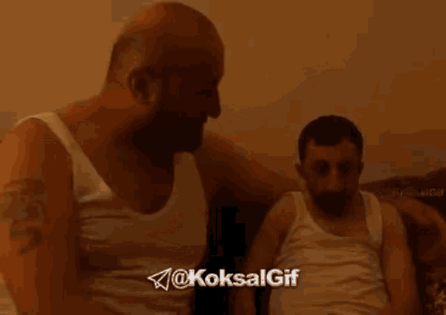 a man in a white tank top is sitting on a couch kissing another man .