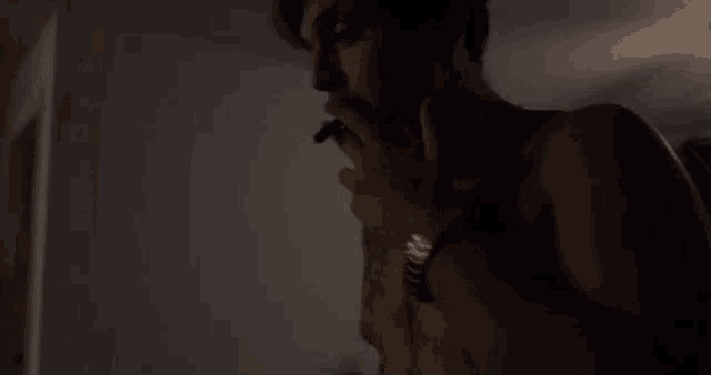 a shirtless man is smoking a cigarette in a dark room while wearing a watch .
