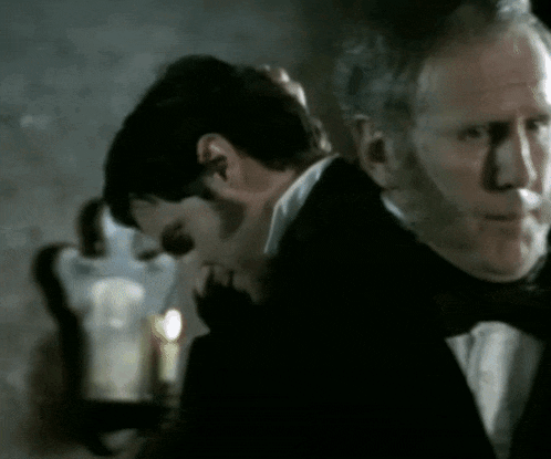 a man in a tuxedo is standing next to another man with a candle in the background