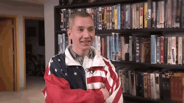 a man wearing an american flag jacket stands in front of a shelf full of dvds including one called a few weeks ago