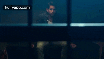 a man is sitting in a dark room looking out of a window with a kulfyapp.com logo in the corner .